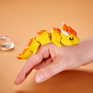 May include: A yellow articulated dragon toy with orange accents and white horns rests on a hand. The dragon has a segmented body and a detailed head. The background is a solid orange color.