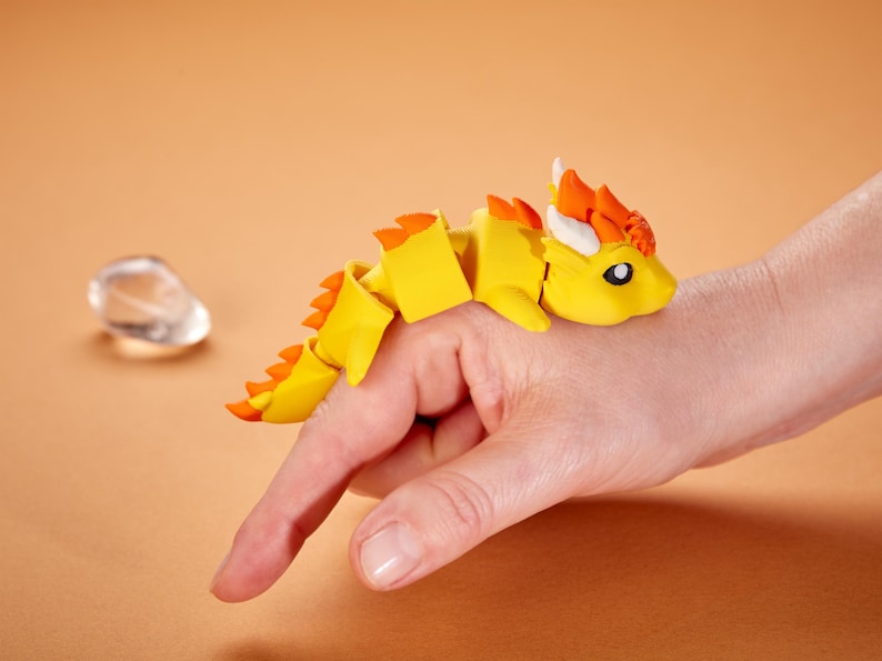 May include: A yellow articulated dragon toy with orange accents and white horns rests on a hand. The dragon has a segmented body and a detailed head. The background is a solid orange color.