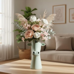May include: A pale green ceramic vase shaped like a cow head, filled with a floral arrangement of light pink roses, white ranunculus, dried pampas grass, and eucalyptus stems. The vase features a smooth matte finish with two small ears and a snout.