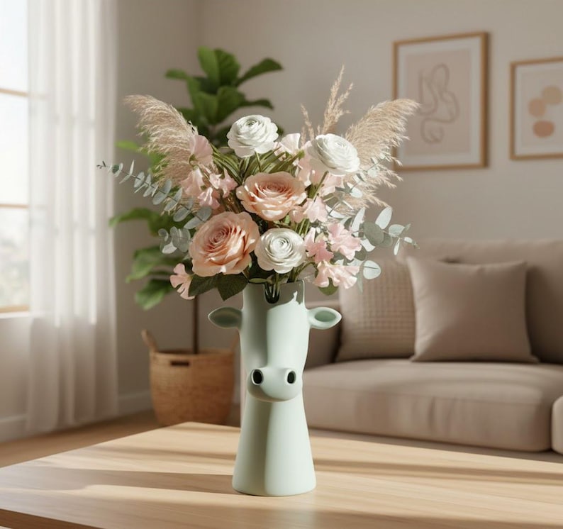 May include: A pale green ceramic vase shaped like a cow head, filled with a floral arrangement of light pink roses, white ranunculus, dried pampas grass, and eucalyptus stems. The vase features a smooth matte finish with two small ears and a snout.