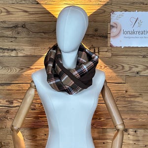 Op de afbeelding: Een bruine en geruite infinity sjaal op een mannequin. De sjaal heeft een mix van bruine, beige, rode en zwarte ruitpatronen. De mannequin staat tegen een houten achtergrond, met een bordje "lonakreativ".
