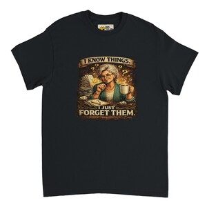 Può includere: T-shirt nero con grafica umoristica. La grafica raffigura una donna con gli occhiali, che tiene una tazza, con la scritta "I KNOW THINGS. I JUST FORGET THEM." Il design include libri, punti interrogativi e una tazza di caffè.