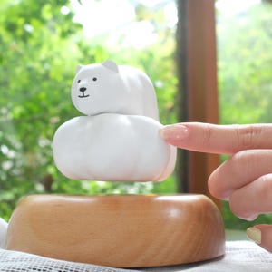 May include: A white polar bear figurine with black eyes and nose sits atop a white cloud-shaped base. The bear is above a wooden bowl, creating a whimsical, decorative piece. The background is a blurred view of green foliage.