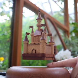 May include: A wooden castle model with a light brown exterior and dark brown accents, including the roof and base. The castle features multiple turrets with small flags and is suspended above a wooden base. The model is intricately detailed and appears to be handcrafted.