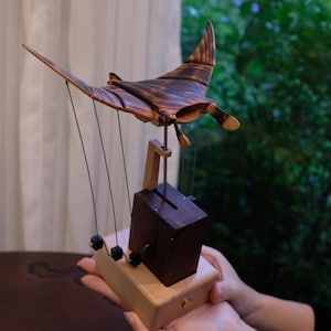 May include: A handcrafted wooden sculpture of a manta ray, featuring a dark brown finish with lighter wood accents. The sculpture is suspended by thin wires from a wooden frame and base. The wingspan appears to be approximately 12 inches.