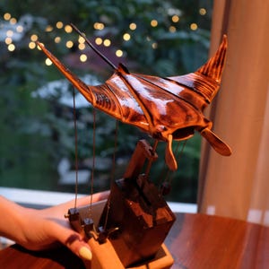 Può includere: Scultura artigianale in legno di manta, con finitura scura e lucida. La scultura è sospesa tramite fili sottili da una base in legno, creando un effetto fluttuante. L'apertura alare è di circa 46 cm. Un'opera d'arte unica.