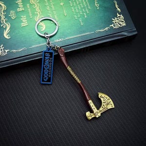 May include: A metal keychain featuring a miniature axe with a gold blade and a brown handle. The keychain also includes a blue tag with the text "GOD OF WAR RAGNAROK". The axe has intricate gold detailing.