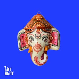 May include: A hand-painted, off-white Ganesh mask with intricate details. The mask features a red and orange headdress, pink ears, and a red spiral trunk. The eyes are outlined in black, and the background is a solid blue.