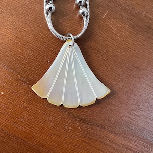 Peut inclure: Un collier argenté avec un pendentif en forme d'éventail. Le pendentif est blanc avec des bords jaunes et des lignes blanches. Le collier a une double chaîne argentée et des perles argentées.