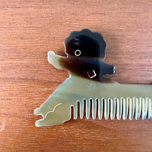 Puede incluir: Un peine vintage con diseño de caniche. El peine está hecho de un material verde claro y marrón, con una silueta de caniche marrón oscuro en la parte superior. Los dientes del peine están espaciados uniformemente y el diseño es caprichoso.