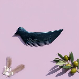 May include: A dark teal bird-shaped comb with the word "FINUE" etched on its back. The comb is set against a light purple background, with olive branches and feathers in the corners. A unique hair care accessory.