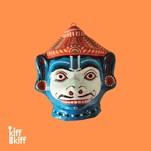May include: A hand-painted, blue and white mask with a smiling face and a red and orange hat. The mask has large eyes, a mustache, and decorative details. The background is a solid orange color.
