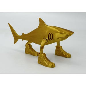 May include: A gold-colored shark figurine with articulated legs and wearing gold high-top sneakers. The shark has a dorsal fin, tail fin, and gills. The figurine is a whimsical decorative item.