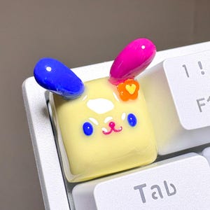 May include: A square yellow keyboard keycap featuring a glossy finish, a cartoon animal face with blue eyes and a pink mouth, one blue ear, one magenta ear, and a small orange flower accent with a yellow heart center attached to the side.