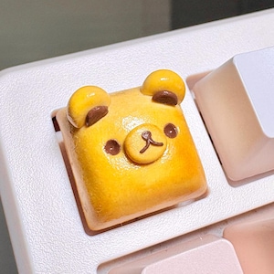 May include: A square mechanical keyboard keycap shaped like a yellow bear face. The keycap features two small rounded ears, two dark brown circular eyes, and a raised snout with a dark brown nose and mouth detail, all with a glossy finish.