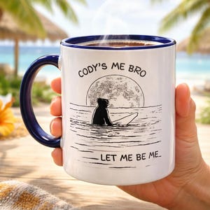 May include: White ceramic mug with a blue handle and rim, featuring the text "Cody's Me Bro" and "Let Me Be Me." The mug has a graphic of a dog surfing at sunset. The mug is filled with a hot beverage.