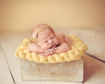 May include: A newborn baby girl sleeping in a yellow knitted basket. The baby is wearing a white headband with a flower detail. The basket is sitting on a light wood surface.