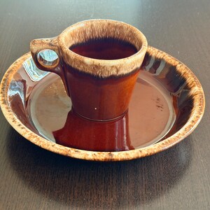 May include: A ceramic cup and saucer set featuring a glossy dark brown glaze with a lighter tan, drip-style rim. The cup has a single handle and a smooth, rounded body, while the matching saucer has a wide, flat base and a raised, textured edge.