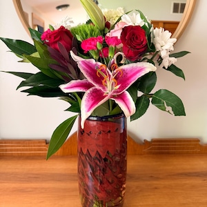 May include: A vibrant floral arrangement in a decorative vase. The bouquet features red roses, pink lilies, carnations, and white flowers, with lush green foliage. The vase is a cylindrical shape with a red and brown pattern.