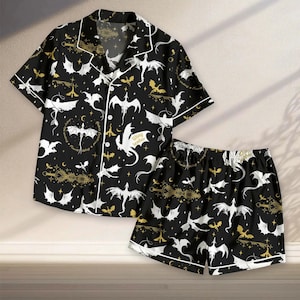 May include: Black pajama set with a dragon pattern. The short-sleeved shirt and shorts feature white and gold dragons, moons, and stars on a black background. The shirt has white trim and buttons. The shorts have an elastic waistband.