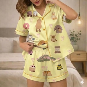 May include: Yellow pajama set with a button-down shirt and shorts. The set is decorated with cartoon illustrations of various food items and animals, including a peanut, duck, and bunny. The edges are trimmed with a gold-colored piping.