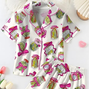 Könnte beinhalten: Weißes Pyjama-Set mit einem Gurkenglas-Muster. Das kurzärmelige Oberteil und die Shorts zeigen Illustrationen von Gurkengläsern mit rosa Schleifen und dem Wort "Pickles" auf Etiketten. Das Set hat rosa Paspeln.