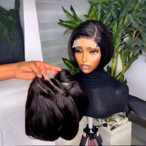 Puede incluir: Una peluca bob negra sobre una cabeza de maniquí. La peluca está peinada con una raya lateral y un corte corto y elegante. El maniquí está sobre un trípode. Una mano sostiene una peluca similar.
