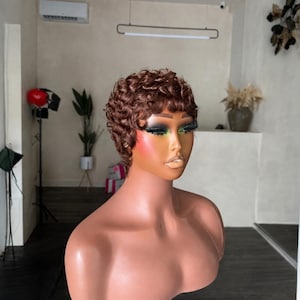 Pode incluir: Uma peruca curta e encaracolada de cor castanha, estilizada com caracóis apertados e texturizados numa cabeça de manequim. A peruca apresenta um tom castanho médio quente com reflexos subtis, proporcionando um aspeto volumoso e em camadas.