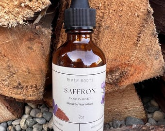 Może przedstawiać: Brązowa szklana butelka z czarnym zakraplaczem. Etykieta głosi "River Roots Saffron Anxiety Relief Organic Saffron Threads 2oz." Butelka jest ustawiona na tle ułożonego drewna opałowego i małych szarych kamieni.