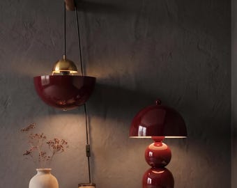 May include: A dark red pendant lamp with a brass dome hangs from a wooden wall hook. Beside it, a dark red table lamp with a spherical base sits on two stacked books. A beige vase, a candle, and a stone tray are also present.