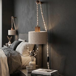 May include: Two beige cylindrical pendant lamps with wooden accents hang from the ceiling. The lamps are suspended by thick, twisted rope cords. The lamps are mounted on a dark gray wall above a bed with gray bedding and pillows. A nightstand with books and vases is below.