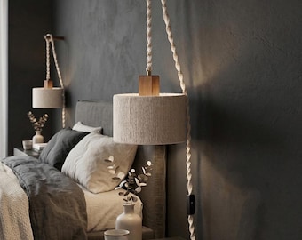 May include: Two beige cylindrical pendant lamps with wooden accents hang from the ceiling. The lamps are suspended by thick, twisted rope cords. The lamps are mounted on a dark gray wall above a bed with gray bedding and pillows. A nightstand with books and vases is below.
