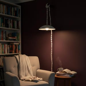 Könnte beinhalten: Eine moderne Pendelleuchte mit weißem Schirm hängt an einem weißen Seil. Die Lampe beleuchtet eine gemütliche Leseecke mit einem beigen Sessel, einer Decke und einem kleinen Beistelltisch mit Büchern und einer Tasse.