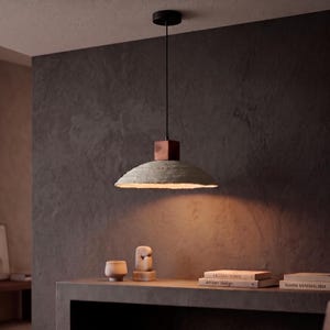 May include: A pendant light fixture featuring a wide, textured, light gray concrete-like shade suspended by a thin black cord from a black ceiling canopy, with a dark brown wooden block connector situated above the shade emitting warm light.