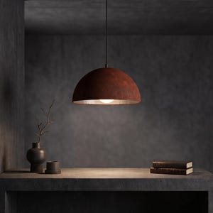May include: A dome-shaped pendant light fixture with a textured, rust-colored exterior and a metallic interior finish. The lamp hangs from a thin black cord and illuminates a single light bulb, casting a warm glow downward from the center.