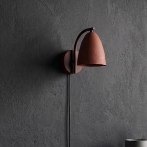 Op de afbeelding: Een wandlamp met een matte, terracotta-kleurige koepelvormige kap en een bijpassende ronde voet. Het armatuur heeft een gebogen arm die de kap met de muurbevestiging verbindt en is voorzien van een dun zwart stroomsnoer.