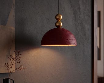 Peut inclure: Une suspension en forme de dôme avec une finition extérieure texturée rouge foncé. Le luminaire présente deux sphères lisses de couleur laiton empilées au sommet, reliées à un fin cordon noir, diffusant une lumière chaude par la base ouverte.