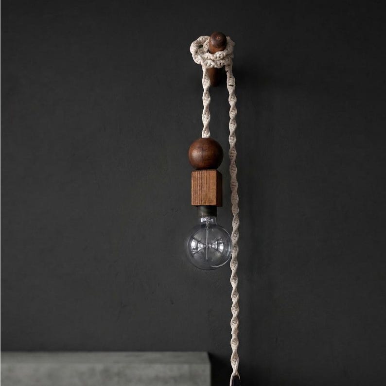 Peut inclure: Un luminaire suspendu moderne avec une ampoule en verre transparent, une sph&egrave;re en bois et un bloc carr&eacute;. La lumi&egrave;re est suspendue &agrave; une corde nou&eacute;e et &agrave; un support en bois. Fond gris fonc&eacute;.
