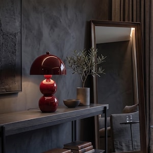 May include: A glossy dark red table lamp with a dome-shaped shade and a base composed of two stacked spheres. The lamp emits a soft glow from the center, positioned on a flat surface next to a small gray bowl and a potted plant in a cylindrical vase.