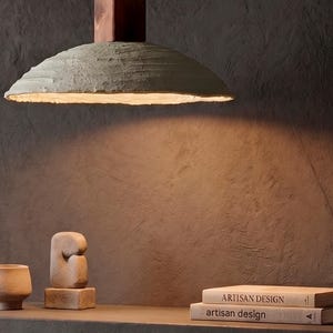May include: A pendant light with a wide, textured, light gray dome shade and a dark brown wooden top mount. The light emits a warm glow from beneath the shade, illuminating the area below where two books titled Artisan Design are stacked.