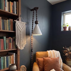 May include: A wall-mounted lamp with a textured white shade and a brown rope cord. The lamp is suspended from a wooden beam. A macrame wall hanging and a bookshelf filled with books are also visible.