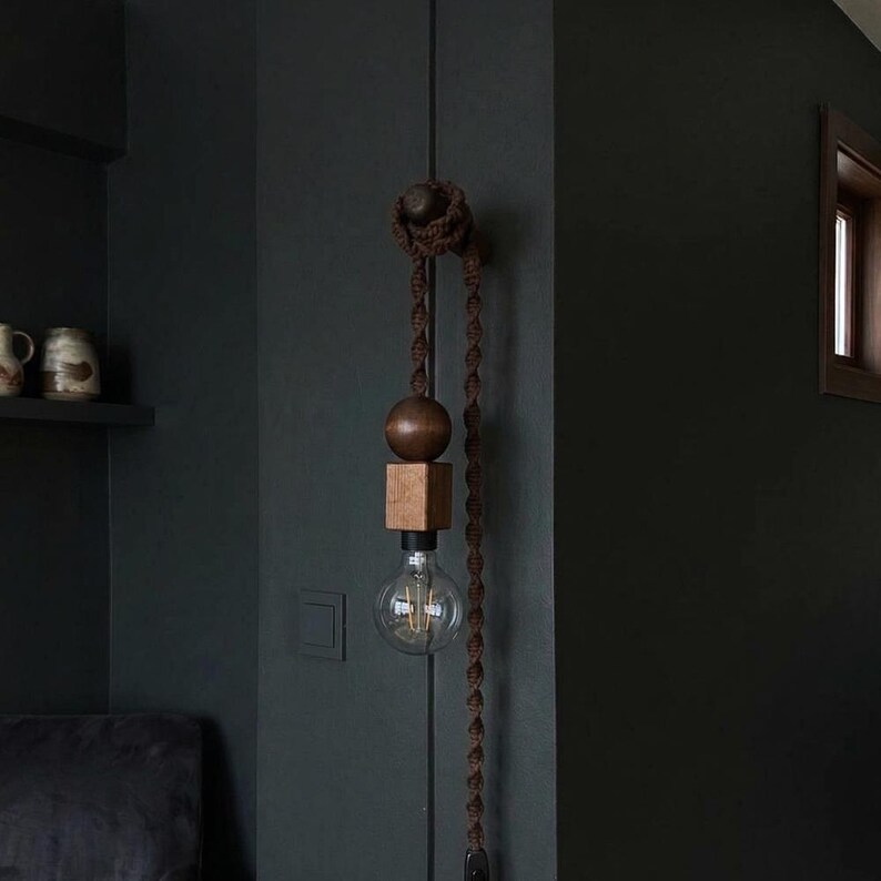 Peut inclure: Un luminaire suspendu d&eacute;coratif avec un design de sph&egrave;re et de cube en bois, suspendu par une corde marron. L'ampoule est visible en dessous des composants en bois. Le luminaire est mont&eacute; sur un mur sombre, cr&eacute;ant une esth&eacute;tique minimaliste.
