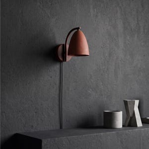 May include: A wall-mounted lamp featuring a matte terracotta-colored dome shade and a curved arm. The fixture is attached to a circular base and includes a thin black power cord extending downward from the light source against the wall.