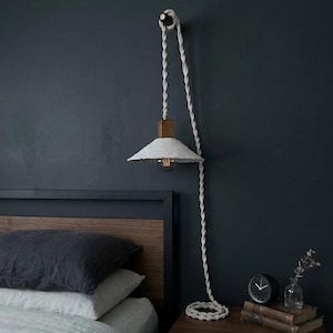May include: A unique wall-mounted lamp with a white paper shade and a wooden block. The lamp hangs from a thick, twisted rope. The design includes a visible lightbulb and is mounted on a dark blue wall. The lamp is next to a bed.