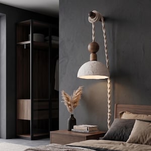 Könnte beinhalten: Ein modernes Schlafzimmer mit einer Hängeleuchte mit strukturiertem, cremefarbenem Schirm und Holzakzenten. Die Leuchte wird von einem dicken Seil gehalten. Ein dunkler Nachttisch beherbergt eine Vase mit Trockenblumen und Büchern. Ein dunkler Kleiderschrank befindet sich im Hintergrund.