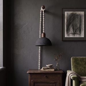 May include: A wall-mounted pendant lamp featuring a dark charcoal dome-shaped shade, a wooden top cap, and a long, thick, cream-colored twisted rope cord that hangs from a wooden wall hook, extending downward past the lamp fixture.