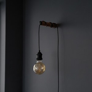 May include: A vintage-style wall-mounted light fixture. It features a wooden bar with a black cord and a single exposed light bulb. The bulb is round and emits a warm, golden glow. The fixture is mounted on a dark gray wall.