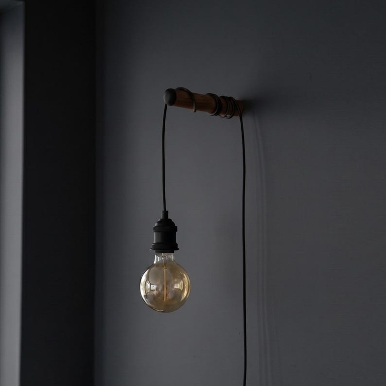 May include: A vintage-style wall-mounted light fixture. It features a wooden bar with a black cord and a single exposed light bulb. The bulb is round and emits a warm, golden glow. The fixture is mounted on a dark gray wall.
