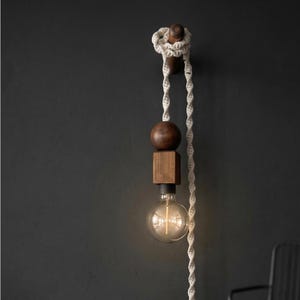 Peut inclure: Une applique murale moderne avec un design en bois et une ampoule visible. Le luminaire comprend une sph&egrave;re et un cube en bois, reli&eacute;s par une corde blanche. L'ampoule est de style Edison, claire et ronde.
