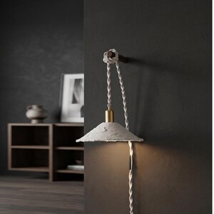 May include: A contemporary wall lamp featuring a textured, off-white shade and a brass-coloured fitting. The lamp is suspended by a twisted white cord, secured to a wooden mount. The design is minimalist, with a neutral colour palette.